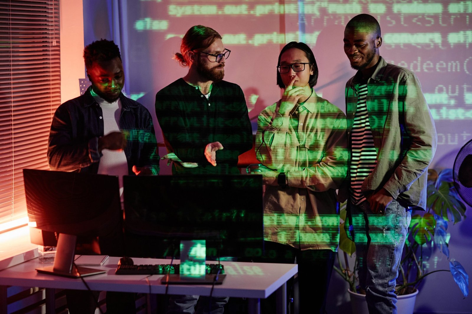 Diverse team of young professionals collaborating around computers with code projected on the wall